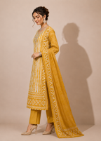 A woman models a traditional Indian outfit consisting of a mustard yellow kurta with white embroidery, matching yellow trousers, and a long dupatta, set against a plain beige background with a soft shadow.