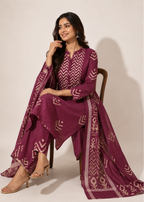 A woman seated on a wooden chair wearing a deep purple traditional outfit with beige geometric patterns and a matching draped scarf, photographed against a plain light beige studio background.