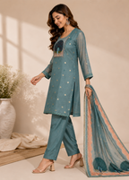 A woman models a teal blue ethnic three-piece suit set consisting of a kurta, pants, and a dupatta, styled in a warm beige room with soft window shadows.