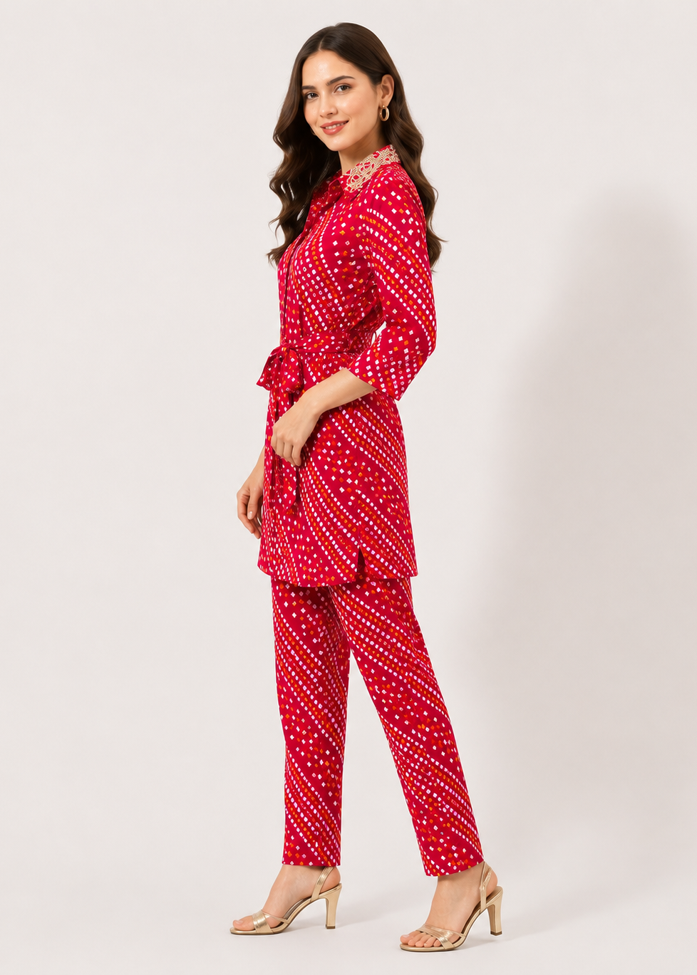 A woman modeling a red two-piece kurta set with a white geometric pattern, consisting of a long-sleeve belted tunic and matching straight-leg pants, paired with clear high-heeled sandals against a light gray studio background.