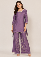 A woman modeling a purple three-piece traditional Indian outfit with a kurta, wide-leg pants, and a matching necklace against a plain beige studio background.