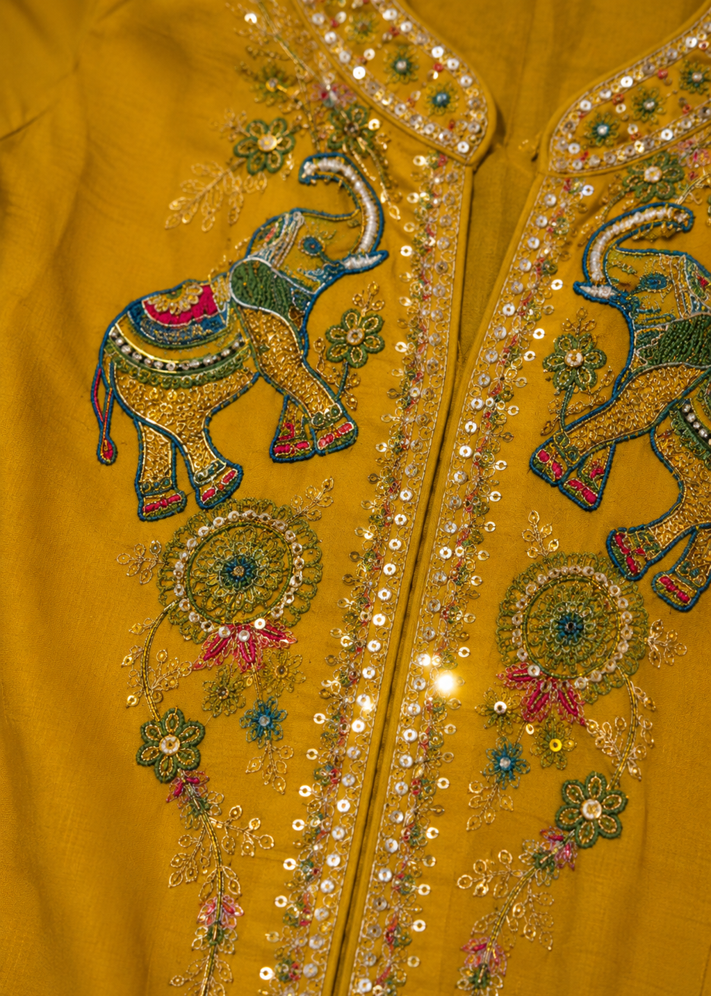 Close-up of a mustard yellow embroidered garment featuring two large elephant motifs and ornate sequin and bead embellishments along the front opening.