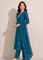 A woman modeling a teal blue traditional Indian outfit consisting of a long kurta with matching pants and a sheer dupatta, decorated with gold embroidery and white pom-pom trim, set against a plain beige studio background.