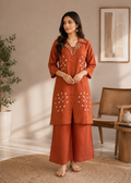Woman wearing a rust-colored embroidered outfit in a room with a plant and wooden furniture.