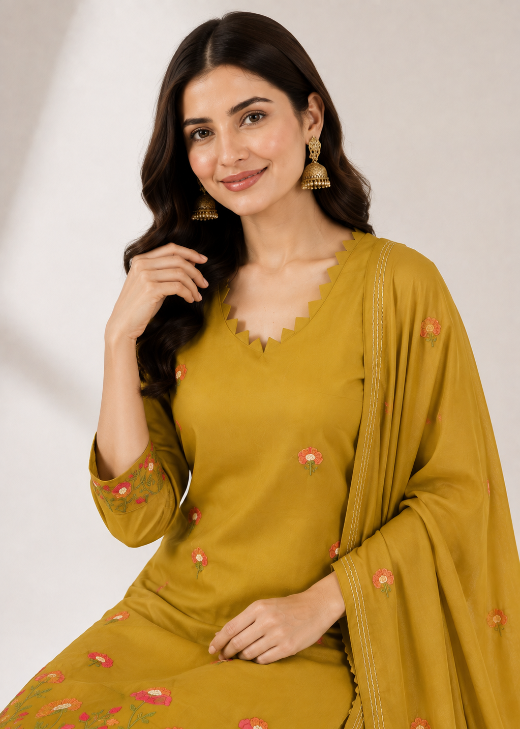 A woman wearing a mustard yellow ethnic suit with pink and green floral embroidery, featuring a V-neck kurta, 3/4 sleeves, and a matching dupatta, posed against a neutral light gray background.