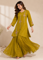 A woman modeling a mustard yellow ethnic kurta set with matching flared pants, featuring pink floral embroidery and silver mirror work details, set against a warm beige background.