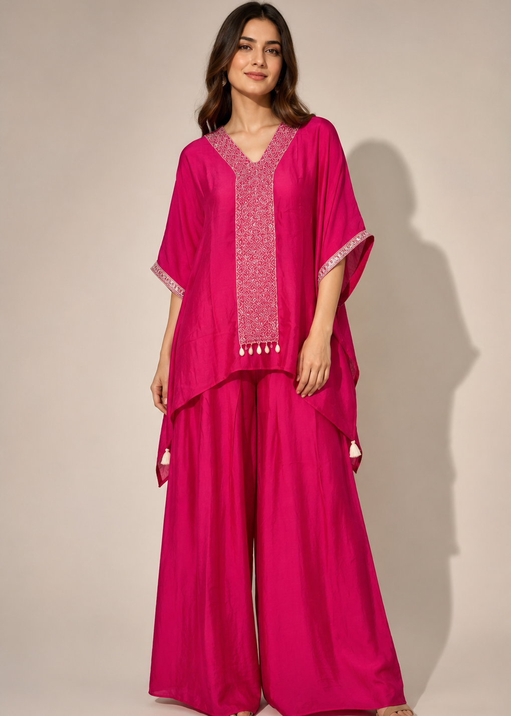 A woman modeling a bright pink traditional outfit consisting of a loose V-neck tunic with embroidered details and matching wide-leg pants, photographed against a plain beige studio background.