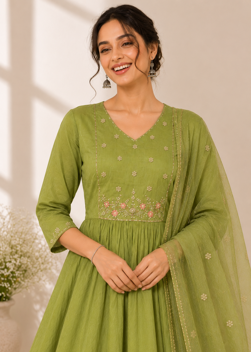 A woman wearing a light olive green traditional Indian outfit with floral embroidery on the bodice and a matching sheer dupatta, standing in a softly lit indoor setting.