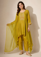 A woman models a three-piece olive green ethnic suit set featuring a long tunic with gold embroidery, matching trousers, and a sheer dupatta, set against a plain beige background.