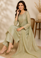 A woman seated on a woven chair wearing a light sage green traditional outfit with a matching sheer draped overlay, shown in an indoor styled setting with a side table and vase in the background.