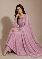 A woman models a traditional Indian outfit consisting of a dusty pink anarkali kurta, matching trousers, and a draped dupatta, styled with layered beaded necklaces and metallic heels against a solid beige studio backdrop.