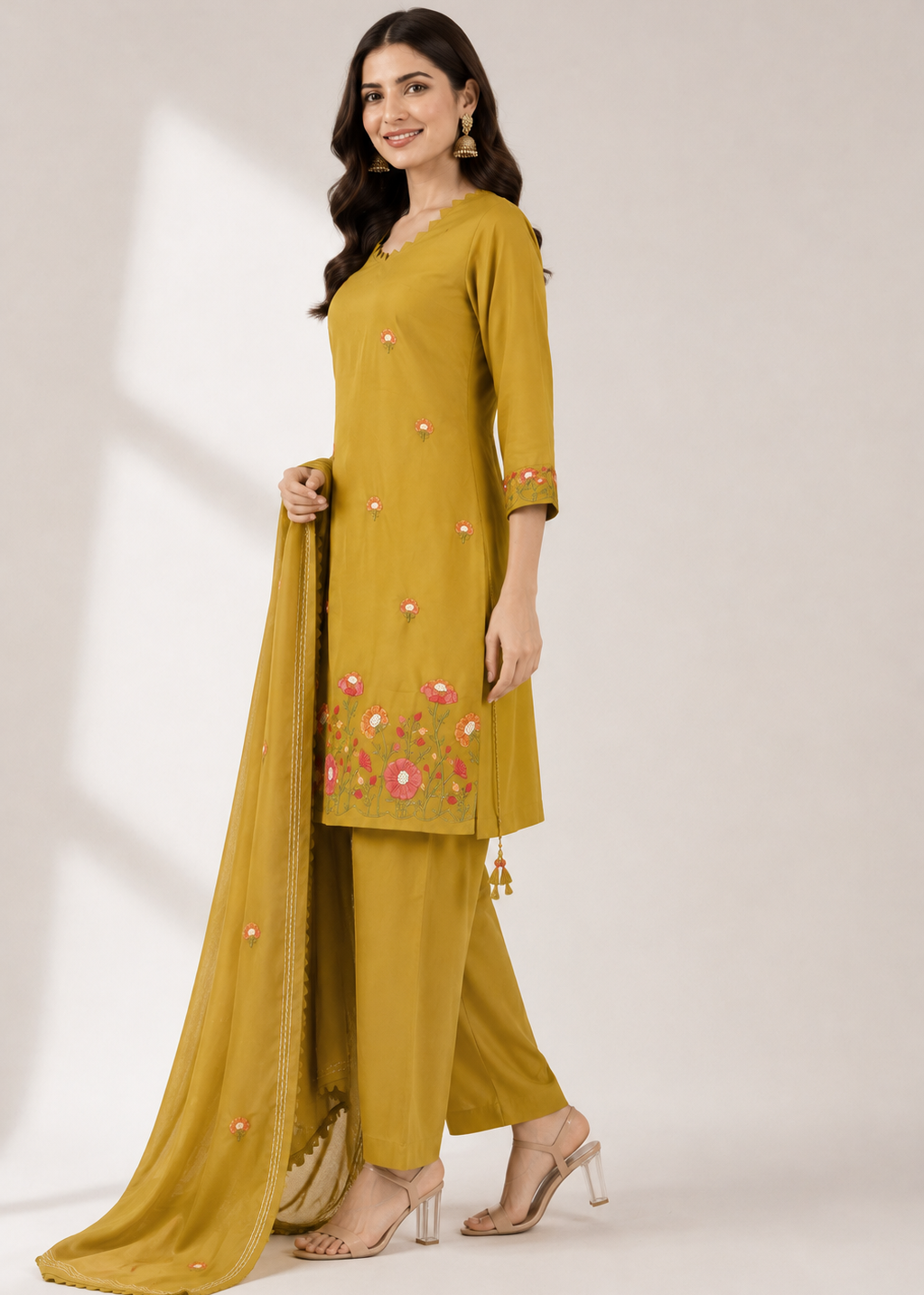 A woman models a mustard yellow salwar kameez suit with pink and white floral embroidery on the kurta and matching dupatta, paired with beige high-heeled sandals against a warm beige studio background.