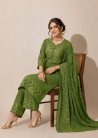 A woman seated on a wooden chair wearing an olive green traditional outfit with white printed embroidery and a matching draped dupatta, photographed against a plain beige studio background.