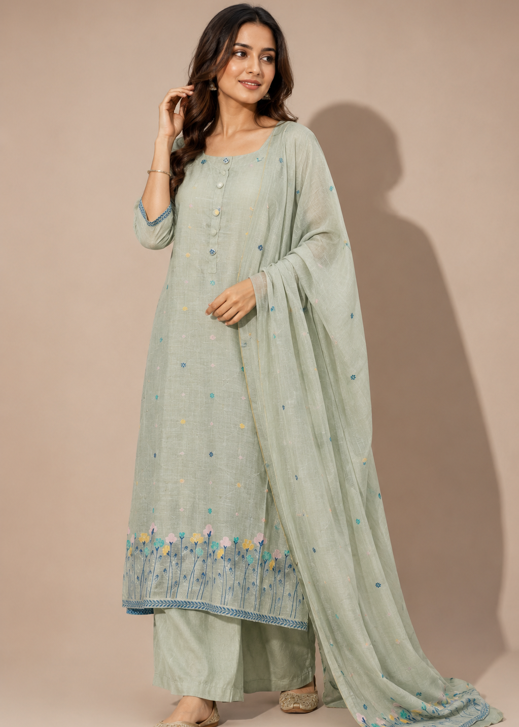A woman modeling a light sage green traditional three-piece outfit with a matching dupatta, featuring embroidered floral borders at the hem and scattered small embroidered motifs, set against a solid beige studio background.