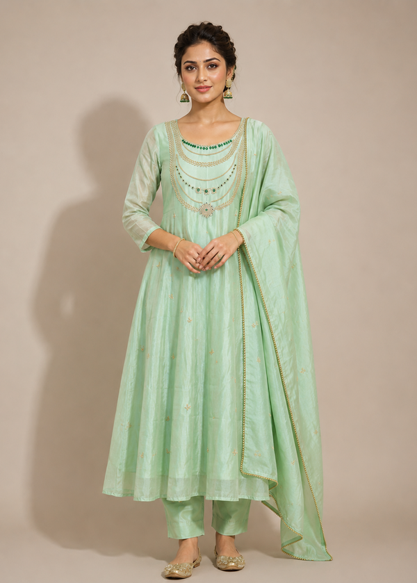 A woman modeling a mint green traditional Indian outfit, featuring a long kurta, matching pants, and a draped dupatta, set against a solid beige studio background.