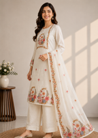 A woman models a traditional Indian salwar kameez set in off-white, featuring intricate floral embroidery in pink, orange, and green. The set includes a long-sleeved tunic, matching wide-leg trousers, and a sheer dupatta draped over her shoulder.