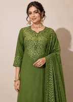 A woman models an olive green traditional Indian salwar kameez suit with intricate white embroidery on the chest and sleeves, paired with a matching embroidered dupatta, set against a plain beige background.