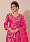A woman modeling a bright pink traditional outfit with a white abstract floral print, ornate silver embroidery around the neckline, and a matching printed dupatta draped over her arm, set against a plain beige studio background.