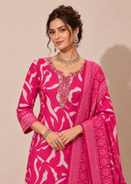A woman modeling a bright pink traditional outfit with a white abstract floral print, ornate silver embroidery around the neckline, and a matching printed dupatta draped over her arm, set against a plain beige studio background.