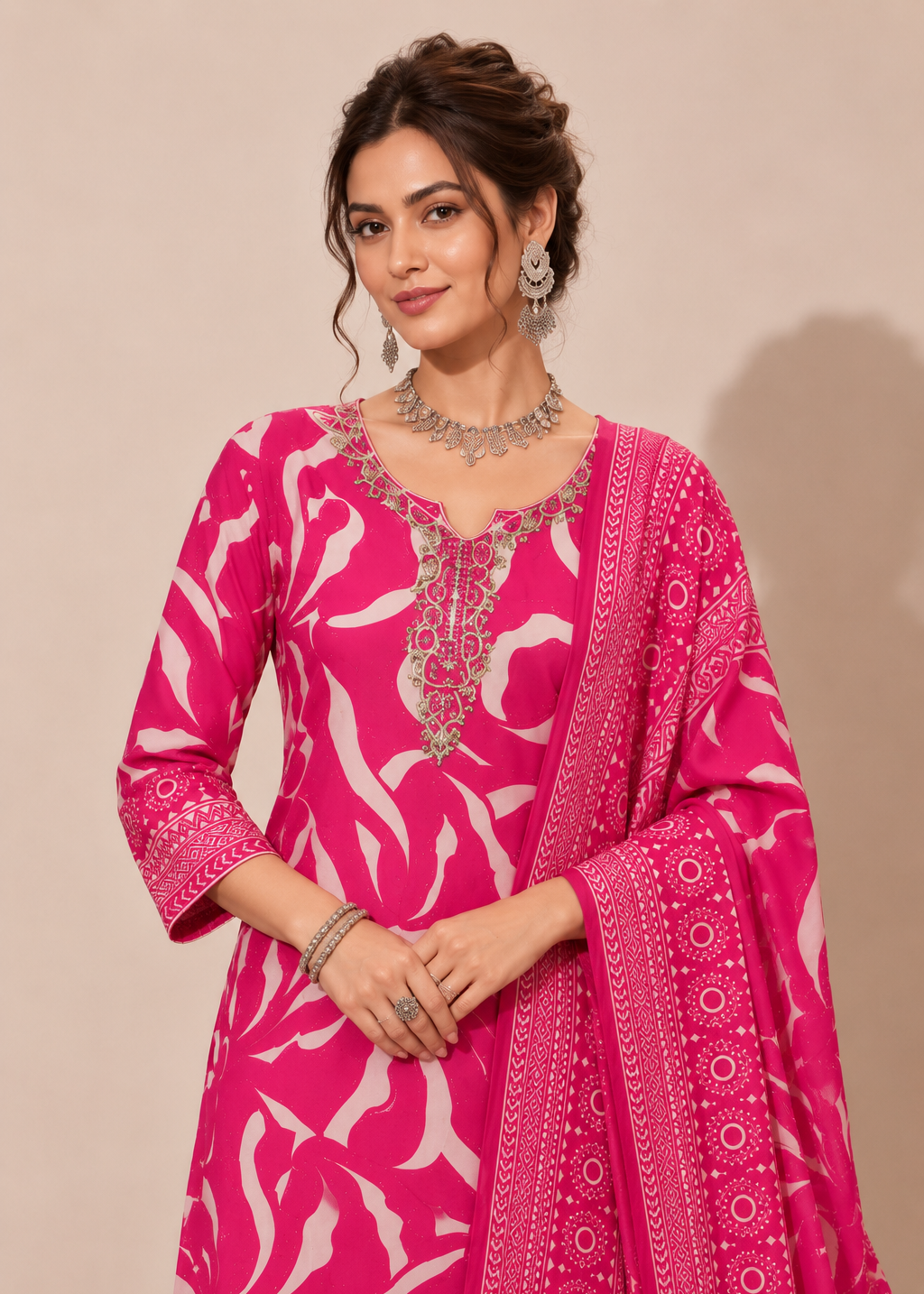 A woman modeling a bright pink traditional outfit with a white abstract floral print, ornate silver embroidery around the neckline, and a matching printed dupatta draped over her arm, set against a plain beige studio background.