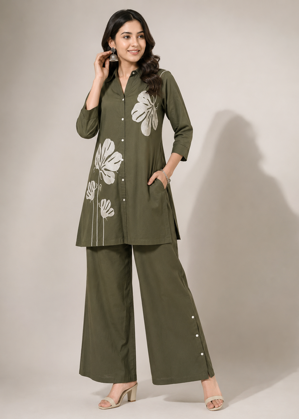 A woman modeling an olive green two-piece kurta set with white floral embroidery, consisting of a long-sleeve button-front tunic and matching wide-leg pants, set against a plain beige studio background.