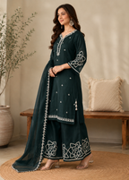 A woman models a dark teal green traditional Indian salwar kameez suit with white floral embroidery and silver mirror work, paired with a matching sheer dupatta, in a warm, neutral-toned indoor setting.
