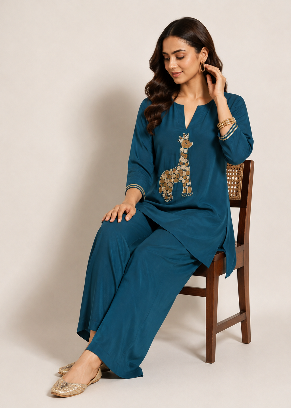A woman seated on a wooden chair wearing a teal blue kurta and matching wide-leg pants set, with a decorative giraffe motif on the chest and gold embellished flat shoes, against a plain beige studio background.