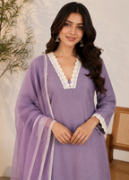 A woman wearing a lavender purple kurta with white lace trim and a matching sheer dupatta, standing in an indoor setting with a neutral wall and a wooden chair partially visible.