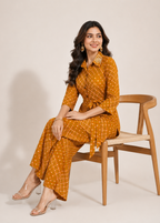 A woman sits on a wooden chair wearing a mustard yellow and white patterned shirtdress with matching wide-leg pants. She is wearing clear high-heeled sandals and gold statement earrings against a plain beige studio background.
