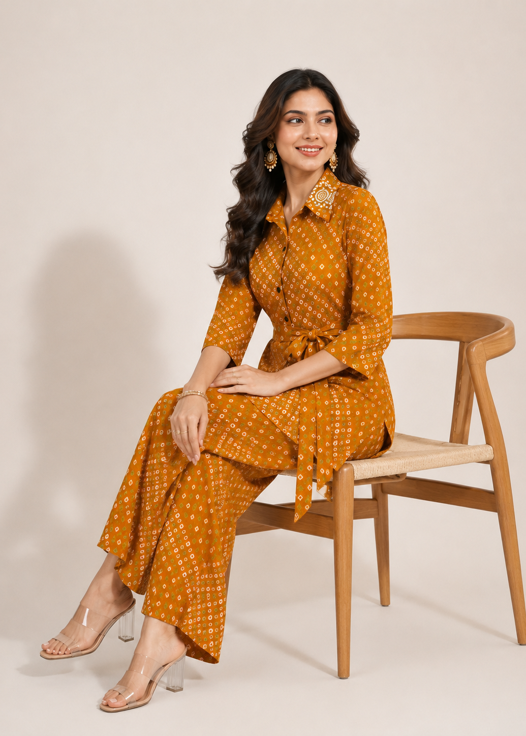 A woman sits on a wooden chair wearing a mustard yellow and white patterned shirtdress with matching wide-leg pants. She is wearing clear high-heeled sandals and gold statement earrings against a plain beige studio background.