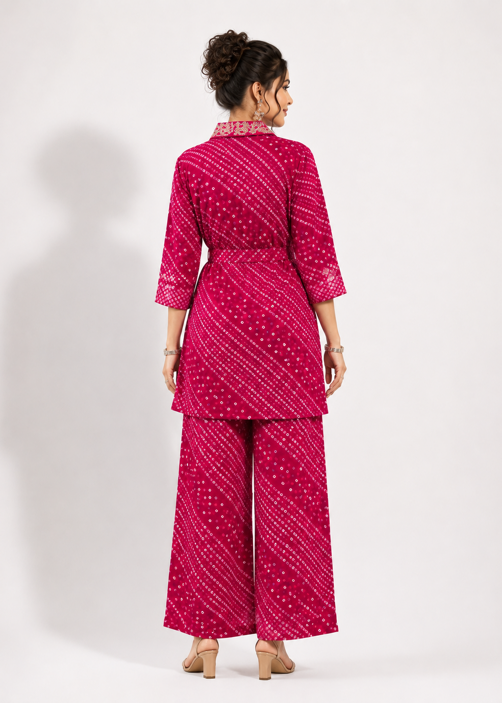 A woman shown from the back wearing a bright pink two-piece outfit with a white bandhani dot pattern, consisting of a short-sleeve tunic top and matching wide-leg pants, set against a plain light gray studio background.