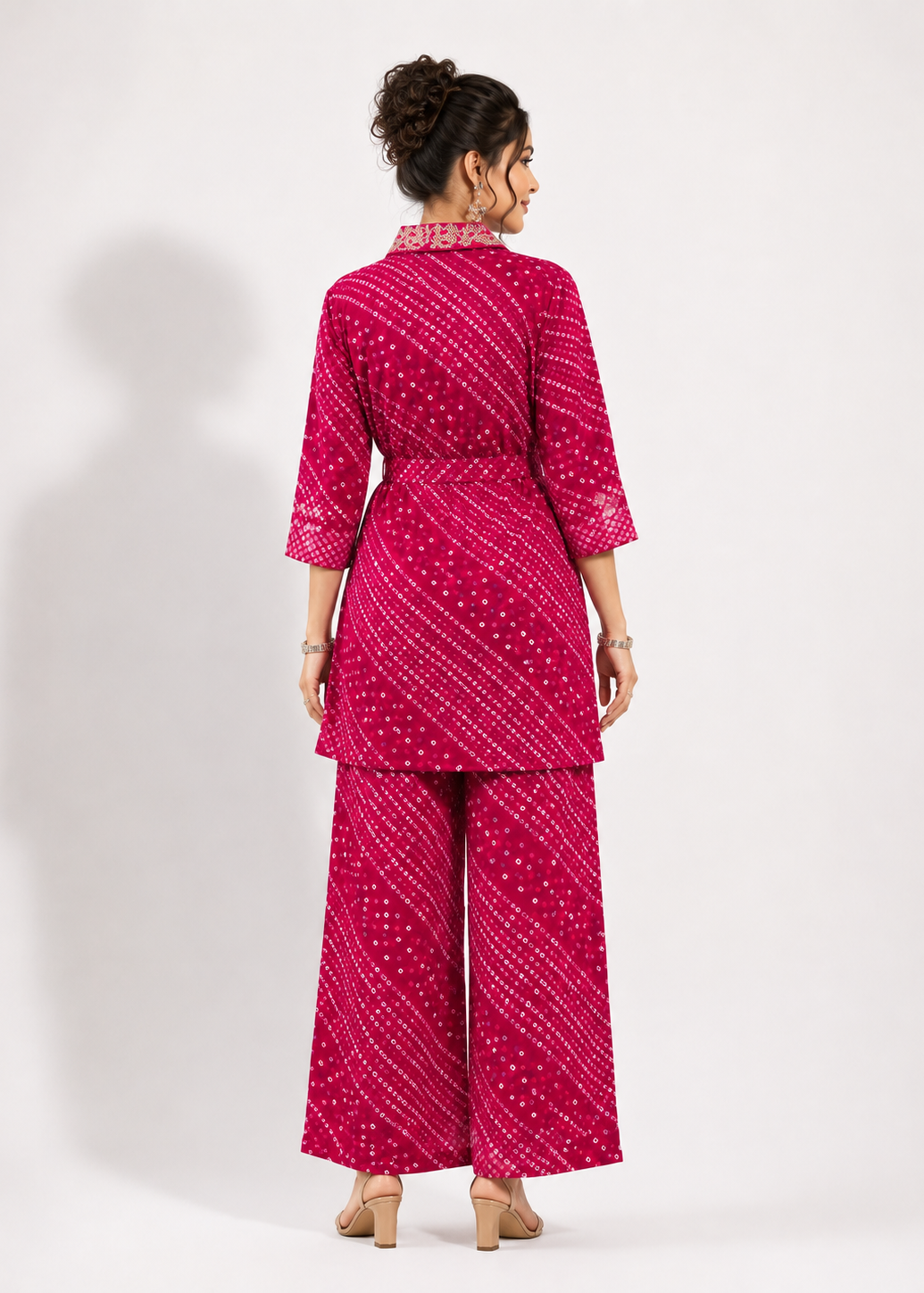A woman shown from the back wearing a bright pink two-piece outfit with a white bandhani dot pattern, consisting of a short-sleeve tunic top and matching wide-leg pants, set against a plain light gray studio background.