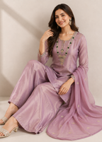 A woman models a lavender purple traditional South Asian outfit consisting of a kurta, wide-leg trousers, and a matching dupatta. The fabric has a subtle sheen and is decorated with gold embroidery on the neckline and scattered motifs on the dupatta.