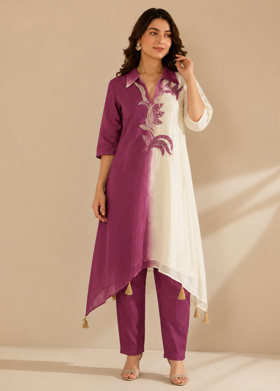A woman models a three-piece traditional Indian outfit consisting of a long-sleeved tunic with a purple-to-white gradient and floral embroidery, matching purple pants, and gold tassel accents, set against a warm beige background.