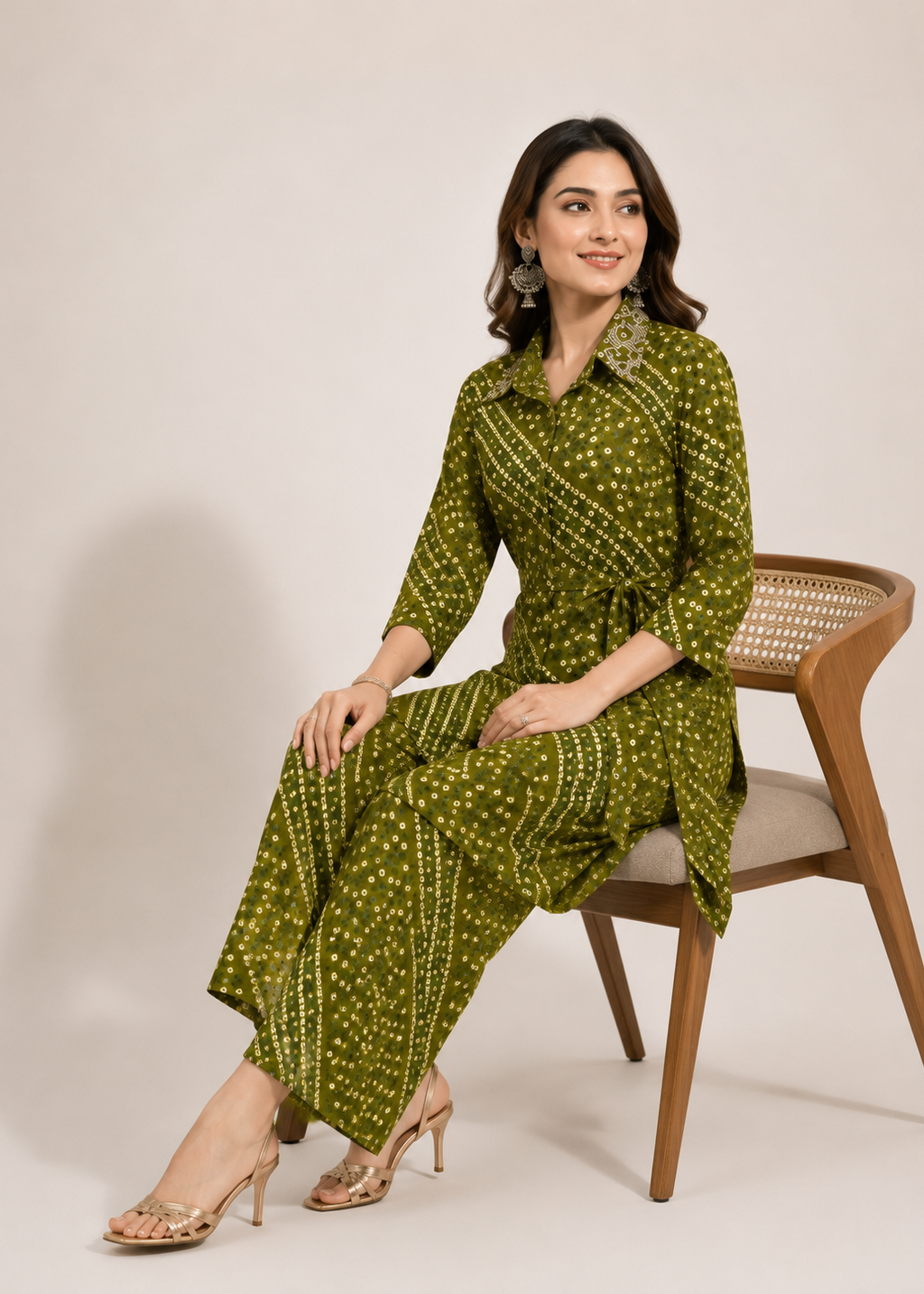 A woman wearing an olive green printed three-piece kurta set sits on a wooden chair against a plain beige studio background. The set includes a long-sleeve collared top, matching pants, and a coordinating belt bag.