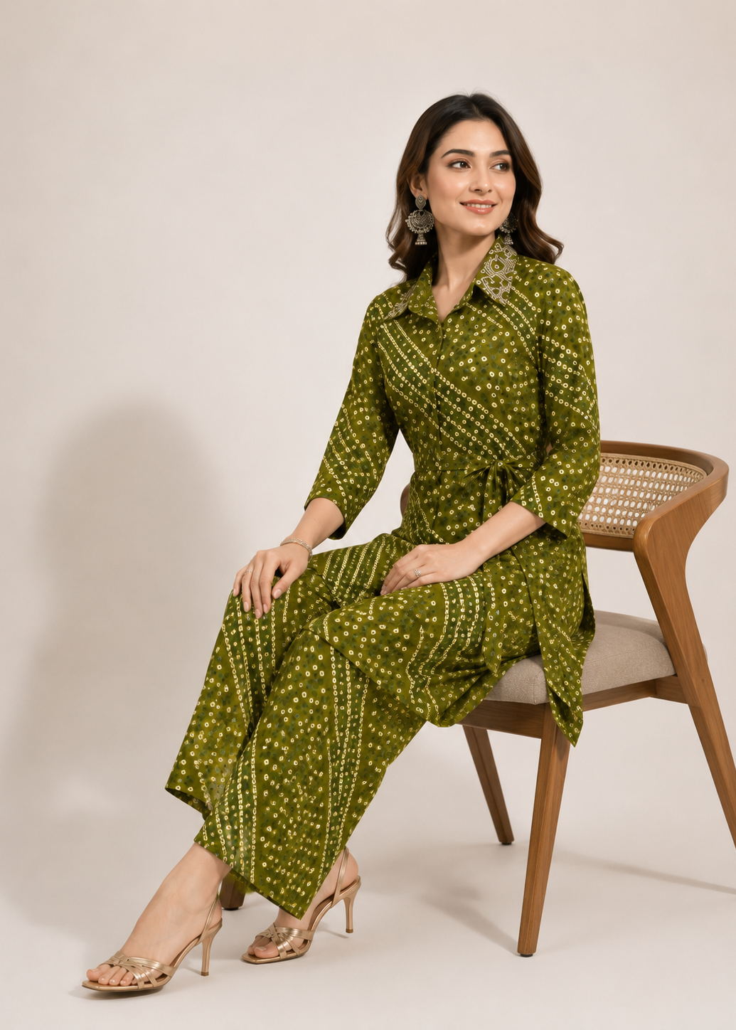 A woman wearing an olive green printed three-piece kurta set sits on a wooden chair against a plain beige studio background. The set includes a long-sleeve collared top, matching pants, and a coordinating belt bag.