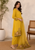 A woman models a yellow three-piece traditional Indian outfit consisting of a kurta, wide-leg pants, and a matching dupatta, decorated with floral and animal embroidery near the hem and cuffs. She is standing in a styled indoor setting with a potted plant and a wooden chair.