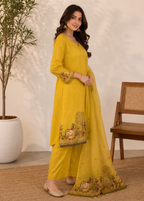 A woman models a yellow three-piece traditional Indian outfit consisting of a kurta, wide-leg pants, and a matching dupatta, decorated with floral and animal embroidery near the hem and cuffs. She is standing in a styled indoor setting with a potted plant and a wooden chair.