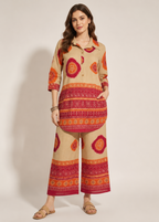 A woman modeling a beige two-piece kurta set with vibrant orange and red bohemian patterns, photographed against a plain light beige studio background.