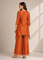 A woman modeling a rust-orange three-piece kurta set with wide-leg pants, shown from the back. The set features white embroidered details on the sleeves and hem, and a decorative tassel fringe.