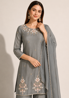 A woman modeling a gray three-piece traditional outfit with floral embroidery on the kurta and dupatta, set against a plain beige background.