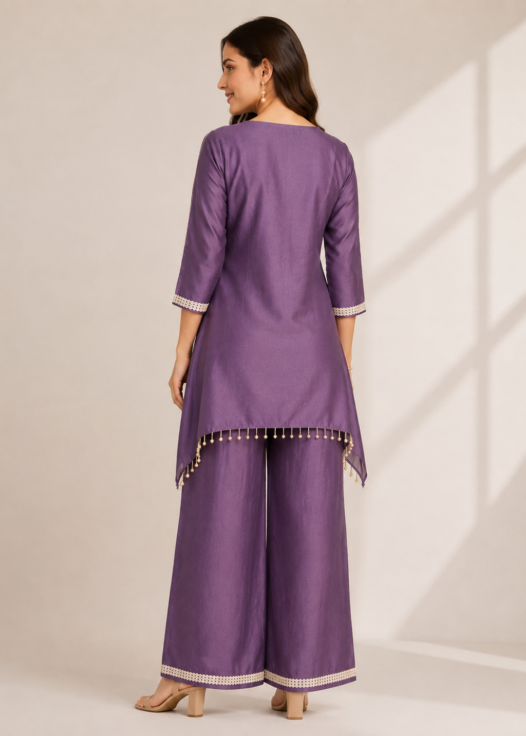 A woman modeling a purple three-piece traditional outfit with a kurta, wide-leg pants, and matching shoes, shown from the back against a warm beige studio background with soft window shadow projections.
