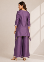A woman modeling a purple three-piece traditional outfit with a kurta, wide-leg pants, and matching shoes, shown from the back against a warm beige studio background with soft window shadow projections.
