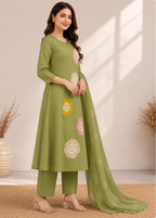A woman models an olive green three-piece salwar kameez set with floral embroidery, consisting of a long tunic, matching trousers, and a dupatta, set against a warm beige background with decorative dried plants.