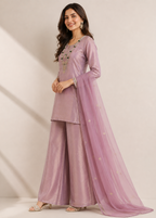 A woman models a lavender purple traditional Indian outfit consisting of a long-sleeve kurta with gold embroidery, matching wide-leg trousers, and a sheer dupatta with small gold star motifs, set against a warm beige studio background.