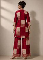 A woman stands with her back to the camera wearing a deep red and beige checkered kurta and wide-leg pants set, photographed in a studio with warm directional lighting and a beige wall background.