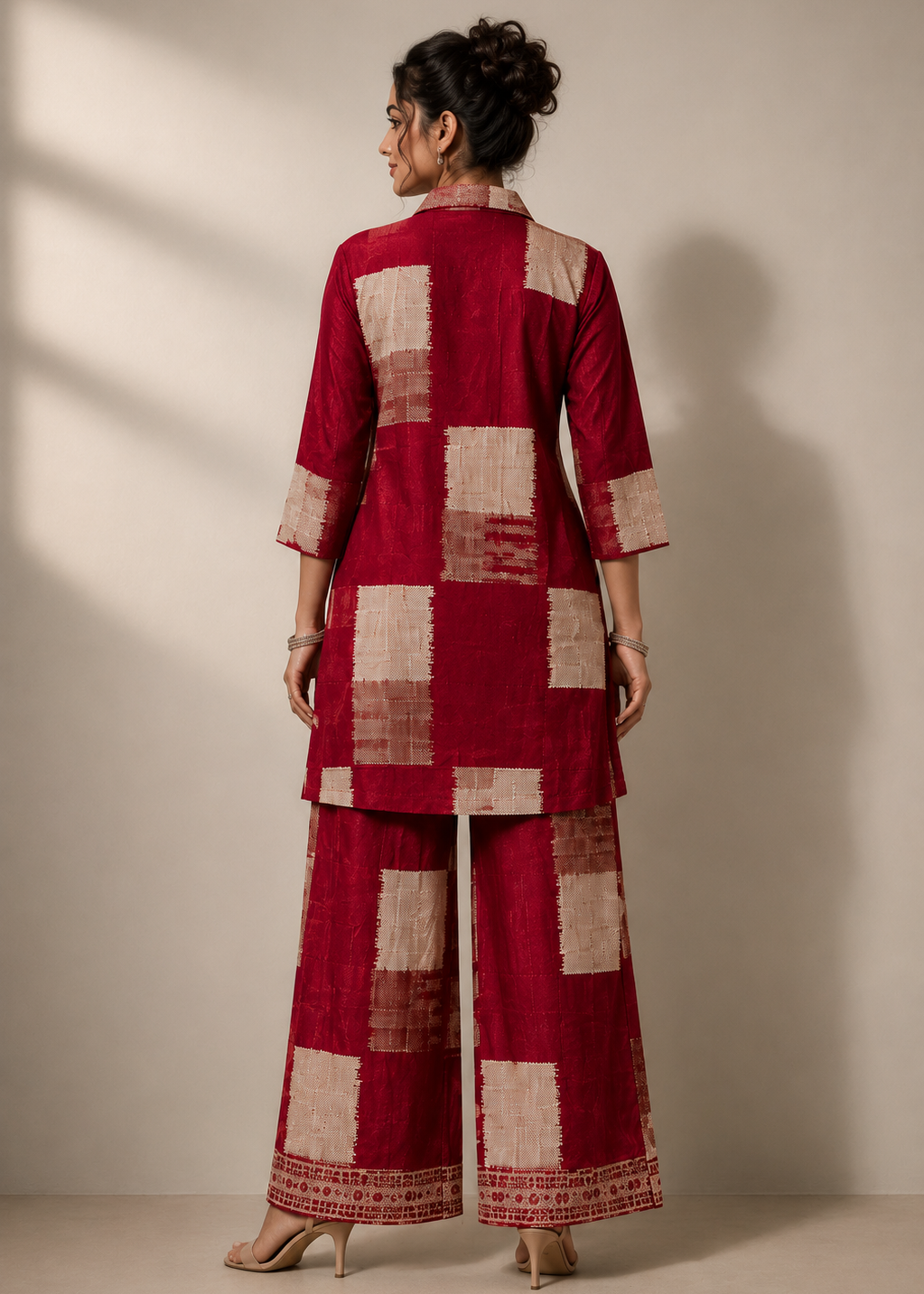 A woman stands with her back to the camera wearing a deep red and beige checkered kurta and wide-leg pants set, photographed in a studio with warm directional lighting and a beige wall background.