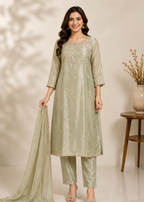 A woman models a light sage green traditional Indian salwar kameez suit with matching dupatta. The kurta features intricate embroidery around the neckline and subtle floral patterns, paired with matching trousers and a sheer dupatta, set against a warm beige background with a wooden side table.