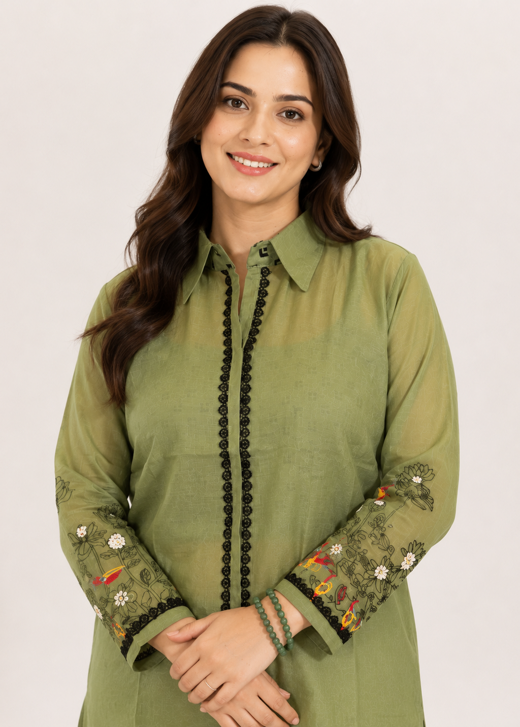A woman wearing a sage green long-sleeve kurta with black beaded trim down the front and floral embroidery on the sleeves, set against a plain light background.