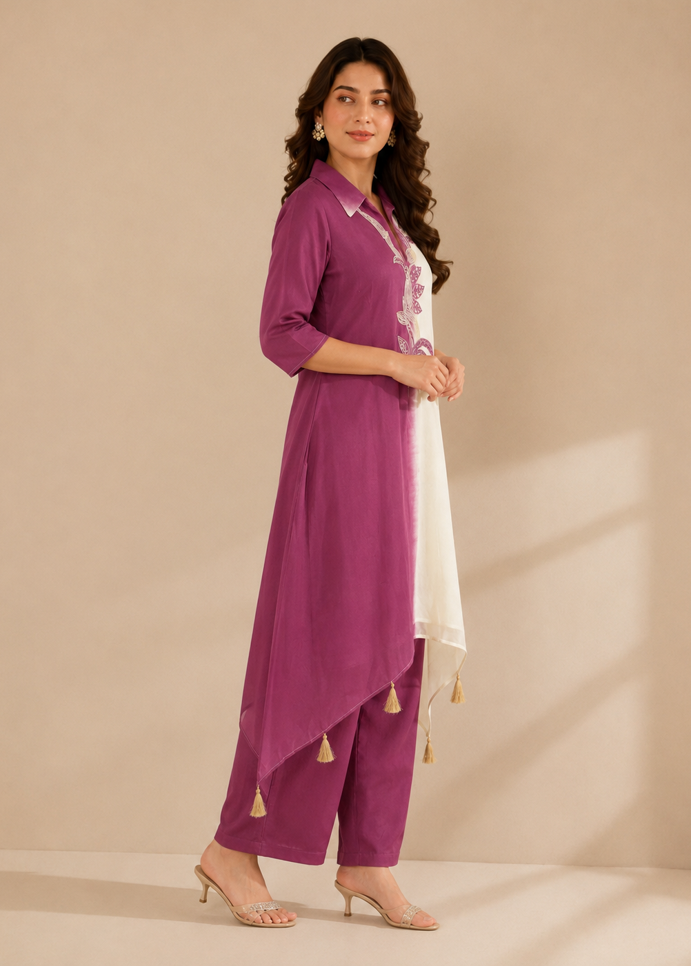 A woman models a three-piece traditional Indian outfit consisting of a purple kurta, matching wide-leg trousers, and a white dupatta with gold tassels, set against a warm beige studio backdrop.